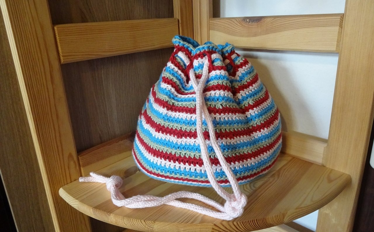 Gehäkelter Kordelzugbeutel, gestreift in Rot, Hellblau, Weiß und Beige auf Holz