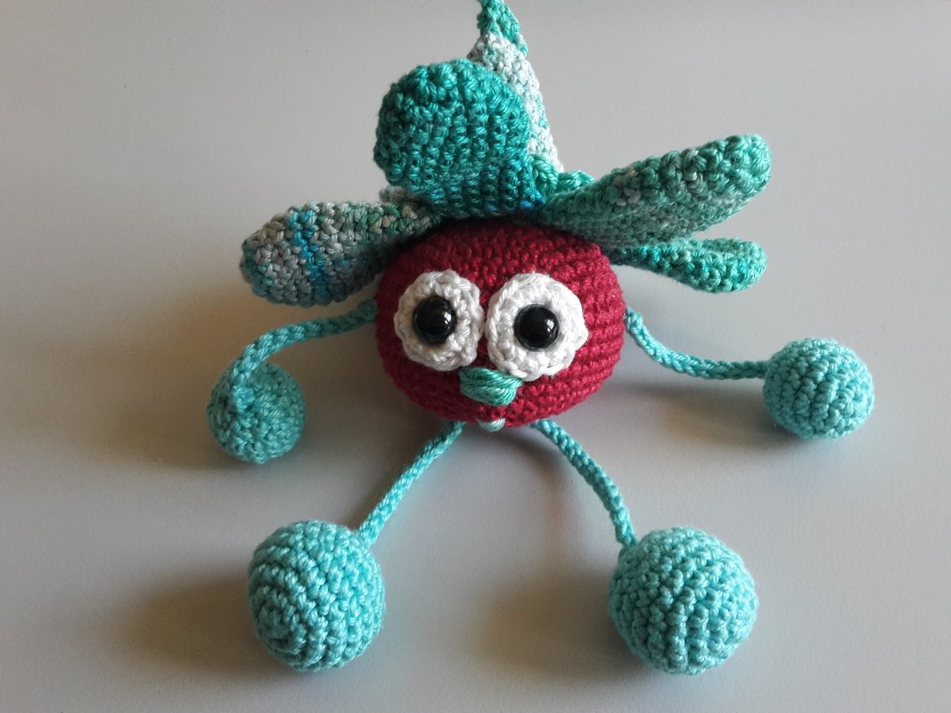Gehäkeltes Amigurumi mit rotem Körper, weißen Augen, türkisfarbenen Blättern und vier Kugelbeinen
