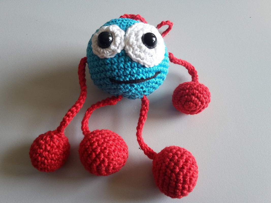 Nahaufnahme eines blauen Amigurumi mit großen Augen und roten Kugel-Gliedmaßen auf hellgrauem Hintergrund.