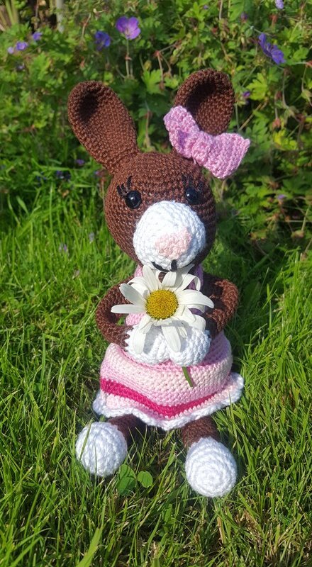 Braunes Amigurumi-Hasenmädchen mit rosa Kleid, Ohrschleife, weißen Pfoten und Margerite im Gras