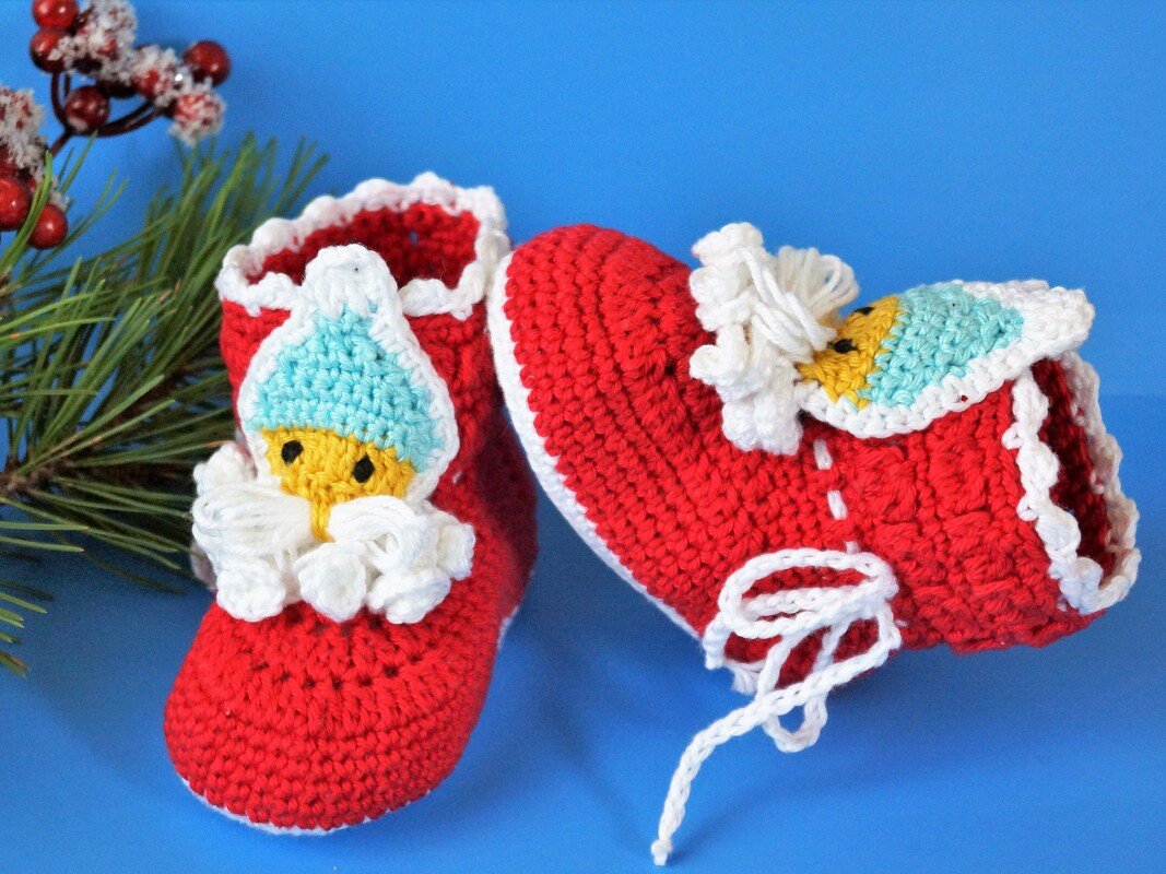 Nahaufnahme zweier roter, gehäkelter Babyschuhe mit Wichtel-Applikationen vor blauem Hintergrund und Tannenzweig