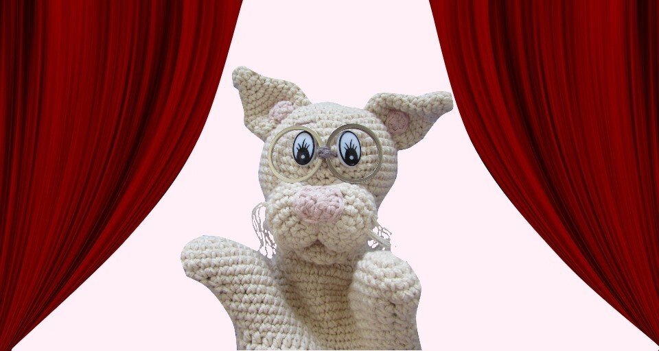Beigefarbene gehäkelte Handpuppe mit Brille vor roten Bühnenvorhängen.