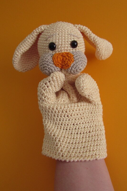 Beige gehäkelte Hasen-Handpuppe mit langen Schlappohren, orangefarbener Nase und grauen Bäckchen auf einer Hand vor orangefarbenem Hintergrund.