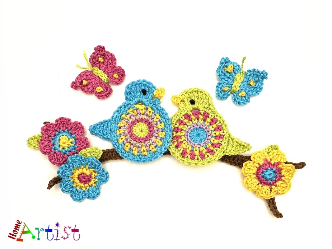Two crocheted bird appliqués—one blue, one yellow—on a brown crocheted branch with colorful crochet flowers and two small crochet butterflies on a white background