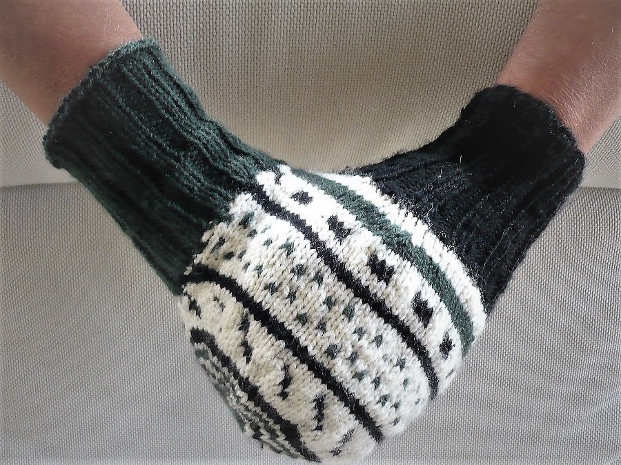 Zwei gestrickte Fausthandschuhe in Dunkelgrün, Creme und Schwarz mit nordischem Muster an Händen getragen.