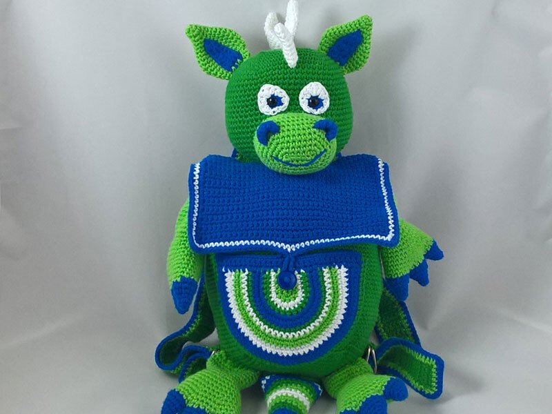 Front view of a green crocheted dragon backpack with a blue flap, white horns, and a patterned front pocket.
