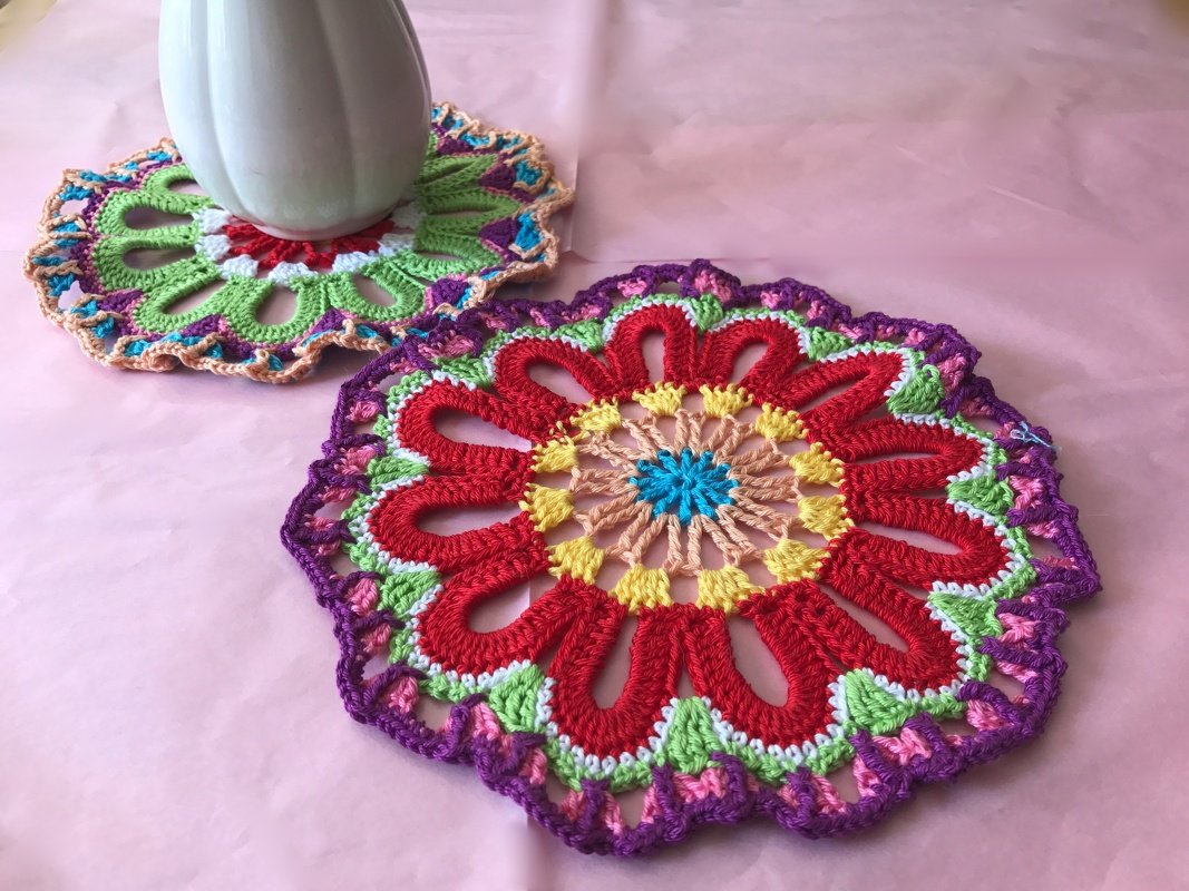 Zwei bunte gehäkelte Deckchen auf rosa Untergrund, eines mit roter Blüte, das andere teilweise von weißer Vase verdeckt