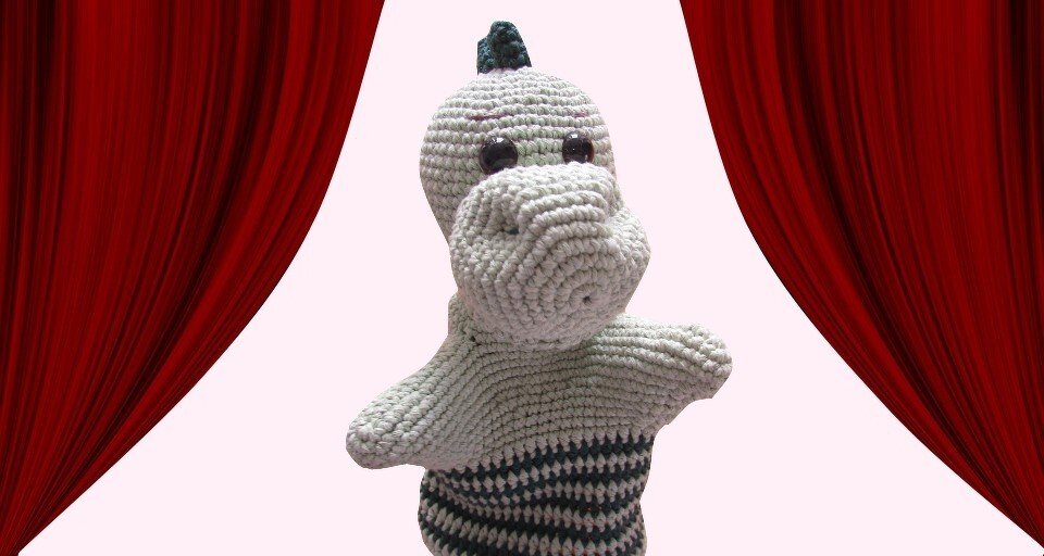 Gehäkelte grün-gestreifte Dino-Handpuppe vor rotem Bühnenvorhang