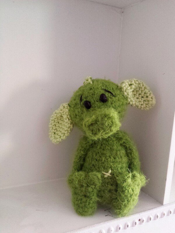 Gehäkelte grüne Amigurumi-Fantasiefigur mit hellgrünen Ohren und schwarzen Knopfaugen auf weißem Regal.