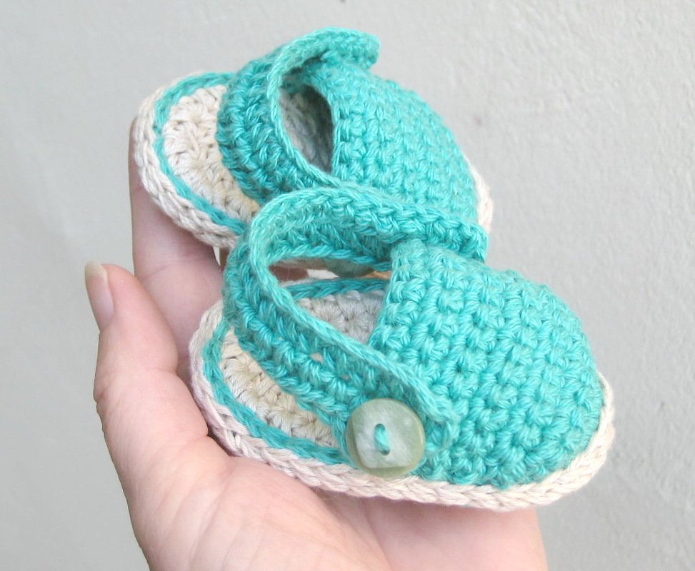 A hand holds two small teal and beige crocheted baby sandals with a button strap.