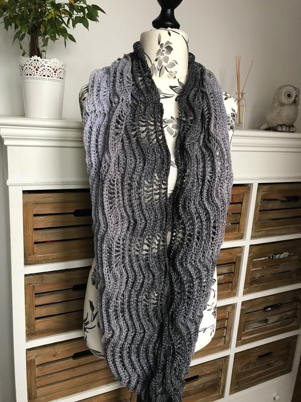 Gehäkelter Loop-Schal in Grautönen mit Farbverlauf und Wellenmuster auf einer Schneiderpuppe drapiert.