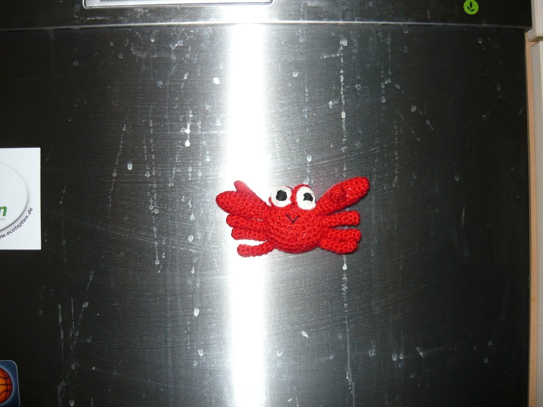 Rote gehäkelte Amigurumi-Krabbe mit großen Augen auf metallischer Oberfläche