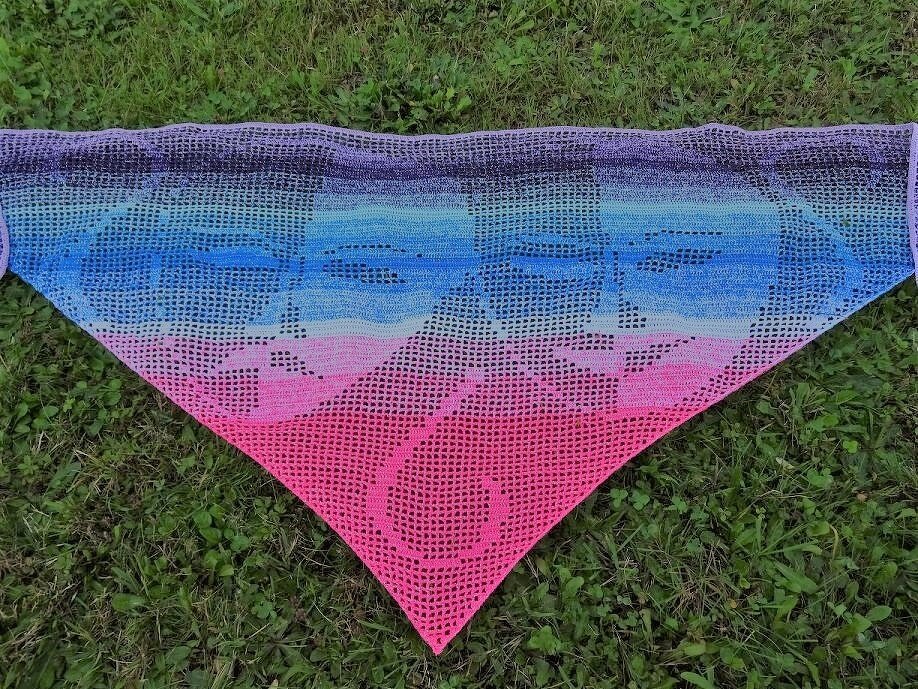 Dreieckiges Filet-Häkeltuch mit stilisierten Gesichtern und lila-blau-pink Farbverlauf auf Gras