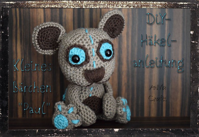 Gehäkelter grauer Teddy „Paul“ mit blauen Akzenten vor dunklem Rahmen