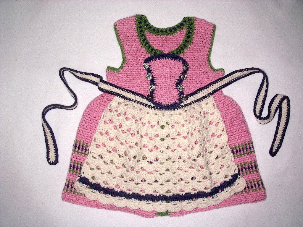 Gehäkeltes rosa Baby-Dirndl mit cremefarbener Schürze sowie grünen und dunkelblauen Details auf weißem Hintergrund