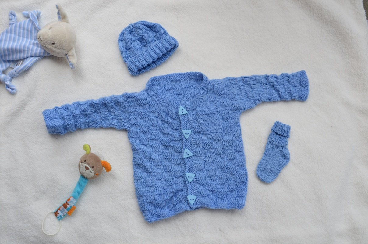 Hellblaues gestricktes Babyjäckchen mit passender Mütze und Socke auf weißer Decke