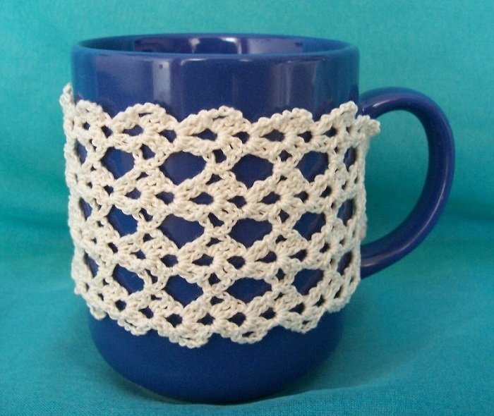 Blaue Tasse mit cremeweißem gehäkeltem Tassenwärmer im Lochmuster vor türkisem Hintergrund.