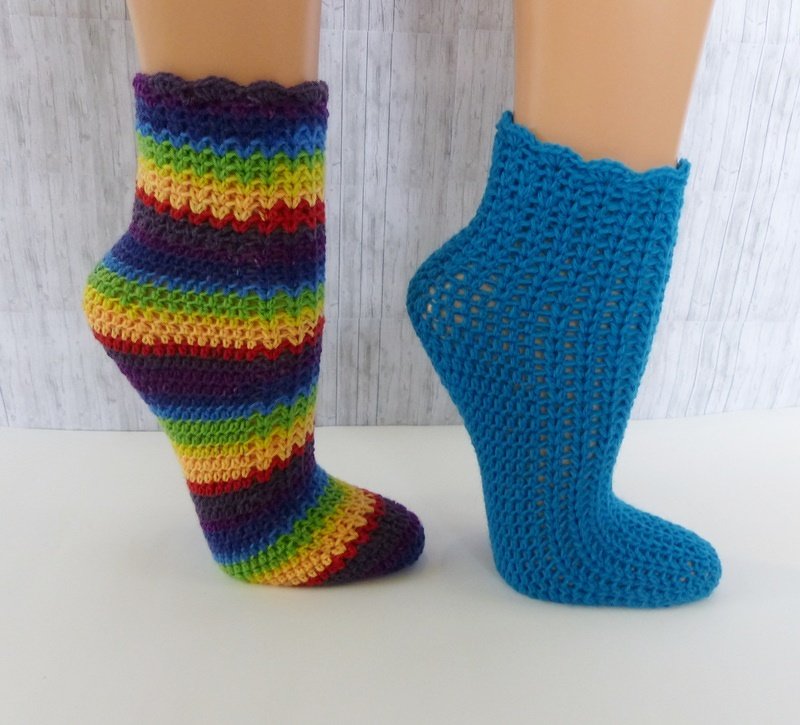Zwei handgehäkelte Socken auf Modellfüßen: gestreifte Regenbogensocke und einfarbige türkise Socke mit Wellenrand