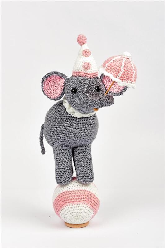 Gehäkelter grauer Amigurumi‑Elefant mit rosa Details auf gestreiftem Ball, hält kleinen Schirm