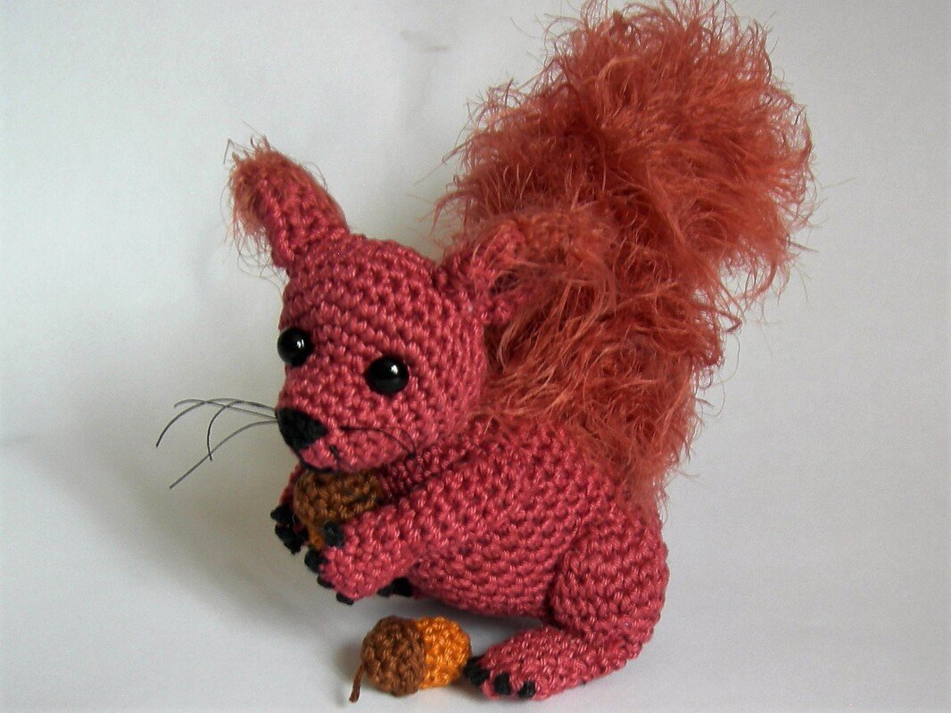 Gehäkeltes rotbraunes Amigurumi-Eichhörnchen mit flauschigem Schwanz und zwei Eicheln vor weißem Hintergrund.