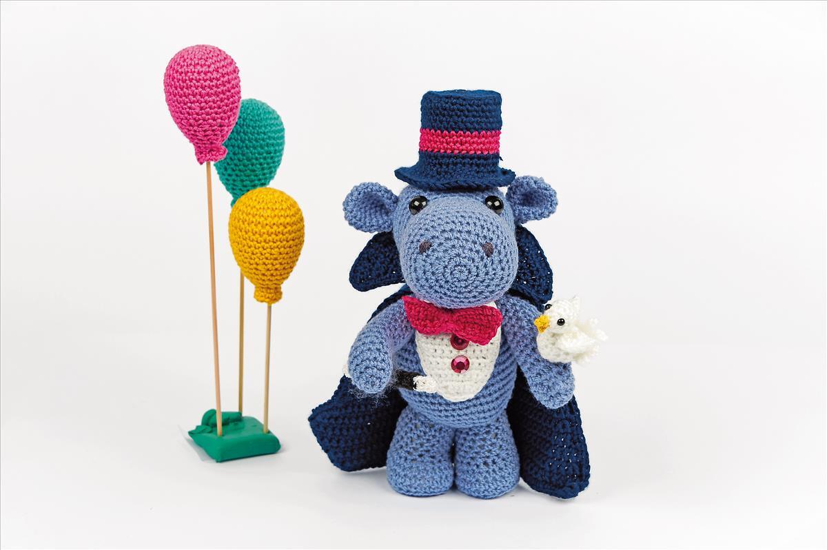 Gehäkeltes blaues Tier-Amigurumi mit Zylinder, Umhang und kleinem weißen Vogel neben bunten Ballons