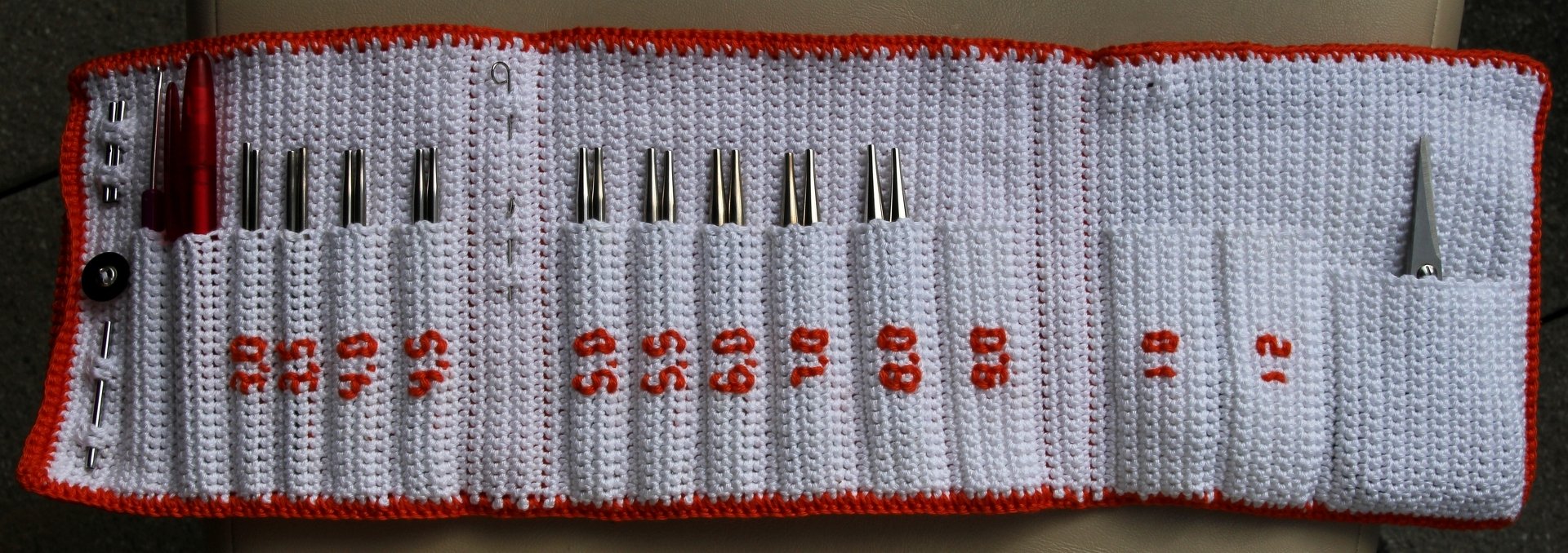 Aufgerolltes weißes Häkel-Etui mit orangefarbenem Rand, gefüllt mit Metall-Stricknadelspitzen und weiterem Strickzubehör.