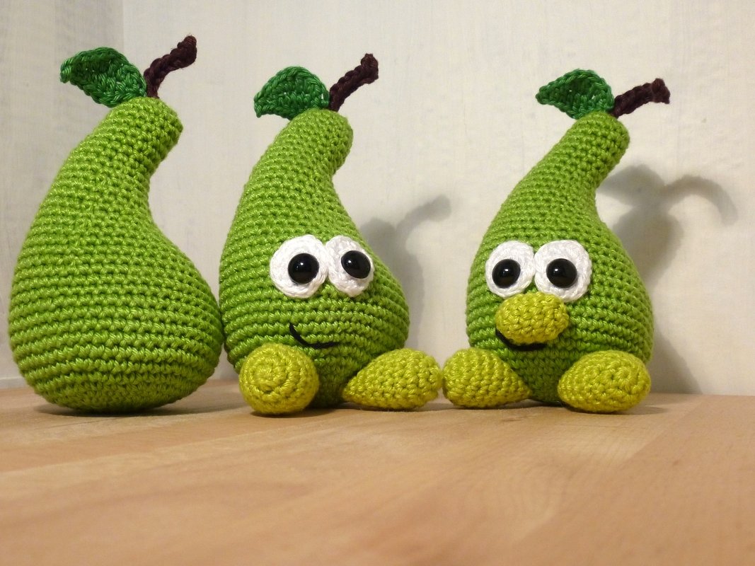 Drei grüne gehäkelte Birnen, zwei mit Gesichtern, auf einer Holzoberfläche