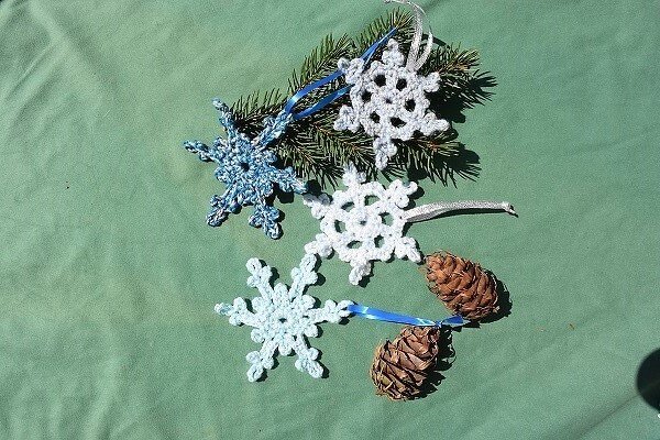 Nahaufnahme gehäkelter Schneeflocken in Weiß und Blau, arrangiert mit Tannenzapfen und einem Tannenzweig auf grünem Stoff.