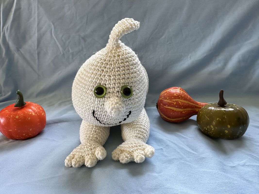 Cremeweißes gehäkeltes Amigurumi-Gespenst mit grünen Augen und drei Zierkürbissen auf hellblauem Stoff.
