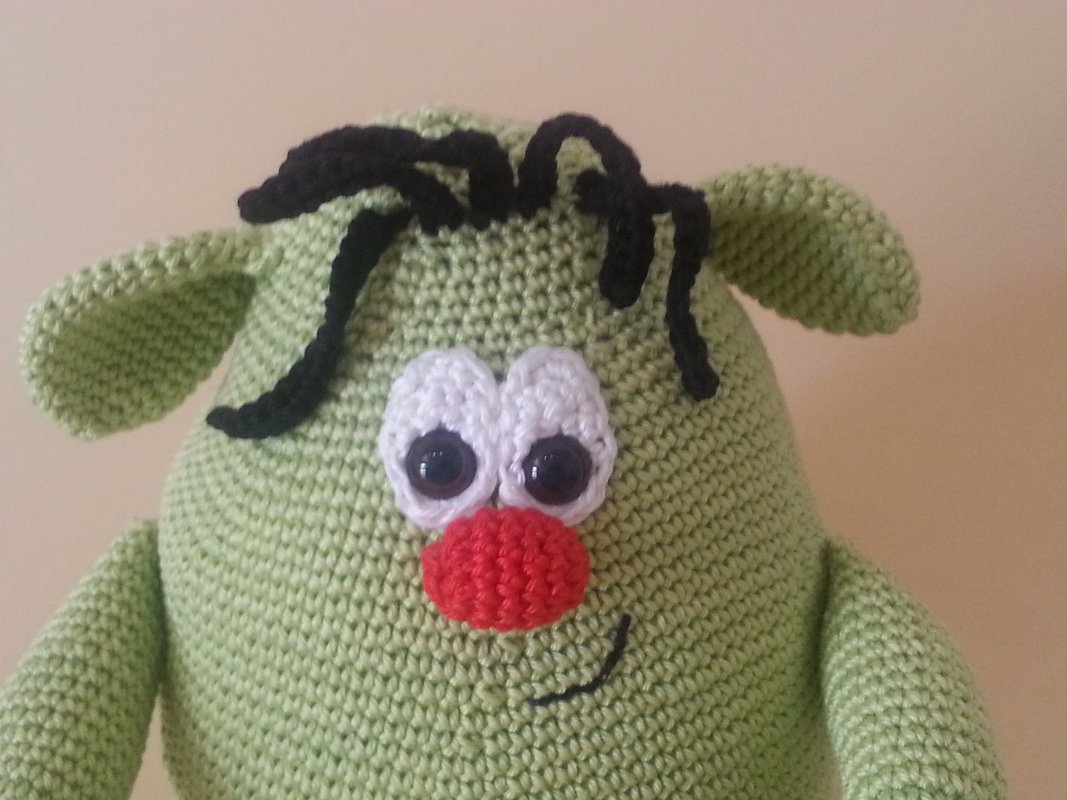 Nahaufnahme eines hellgrünen gehäkelten Amigurumi-Monsters mit großen Augen, roter Nase und schwarzem Garnhaar