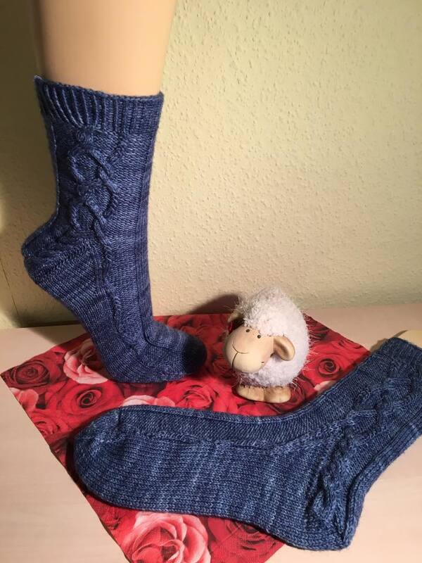 Blaue gestrickte Damensocken mit seitlichem Zopfmuster, eine auf Fußform, eine liegend; kleines Dekorschaf auf Rosenstoff