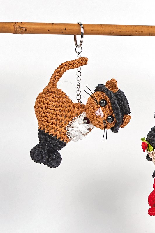 Gehäkelter Katzen-Schlüsselanhänger in Braun und Schwarz mit Spitzenkragen und Mini-Hut, an einem Schlüsselring hängend.