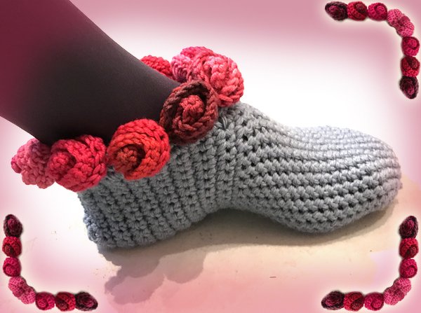Gehäkelter grauer Slipper mit gehäkelter Rosenborte am oberen Rand, getragen an einem dunklen Bein.