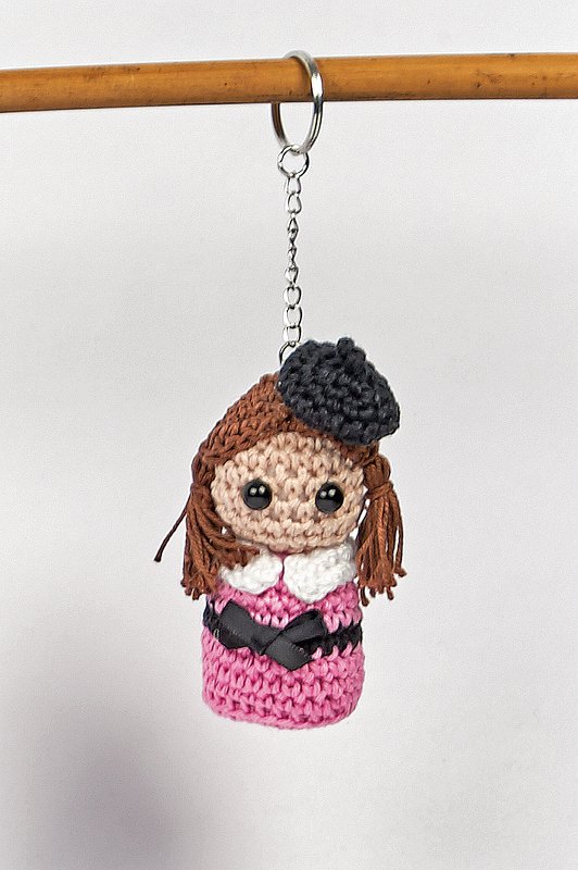 Gehäkelter Amigurumi-Puppen-Schlüsselanhänger mit rosa Kleid, braunen Zöpfen, weißem Kragen und schwarzer Baskenmütze an Metallring und Kette