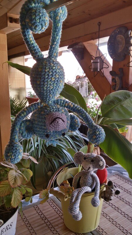 Blau-grün gehäkelter, hängender Amigurumi-Affe und eine graue gehäkelte Maus in einer gelben Gießkanne vor Pflanzen auf einer Veranda.