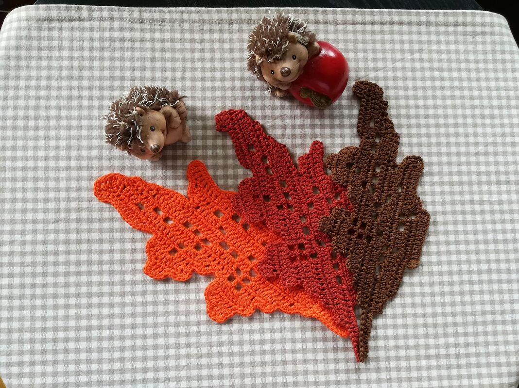 Drei gehäkelte Blätter in Orange, Rot und Braun auf kariertem Stoff neben zwei kleinen Igel‑Figuren.