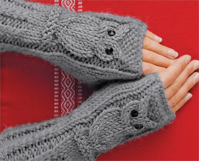 Nahaufnahme grauer, gestrickter Armstulpen mit Zopfmuster und schwarzen Knöpfen an Händen vor rotem Hintergrund.