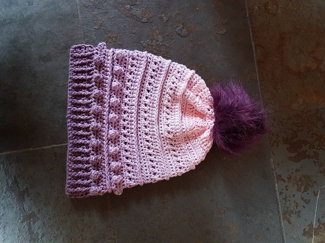 Rosa-lila gehäkelte Beanie mit Rippenbund, Noppenreihen und dunklem flauschigem Bommel auf dunklem Untergrund