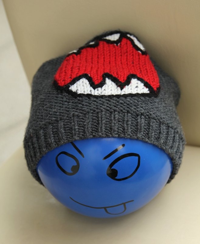 Dunkelgraue gestrickte Beanie mit roter und weißer Zahn‑Applikation auf blauem Ballon als Kopfattrappe