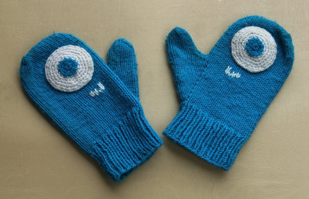 Paar blaues, handgestrickte Kinderfäustlinge mit großen gehäkelten Augen in Weiß und Hellblau.