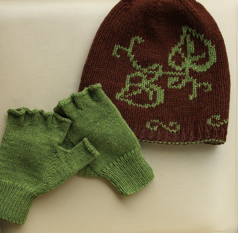 Braune gestrickte Mütze mit grünem Intarsienmuster neben grünen fingerlosen Handschuhen