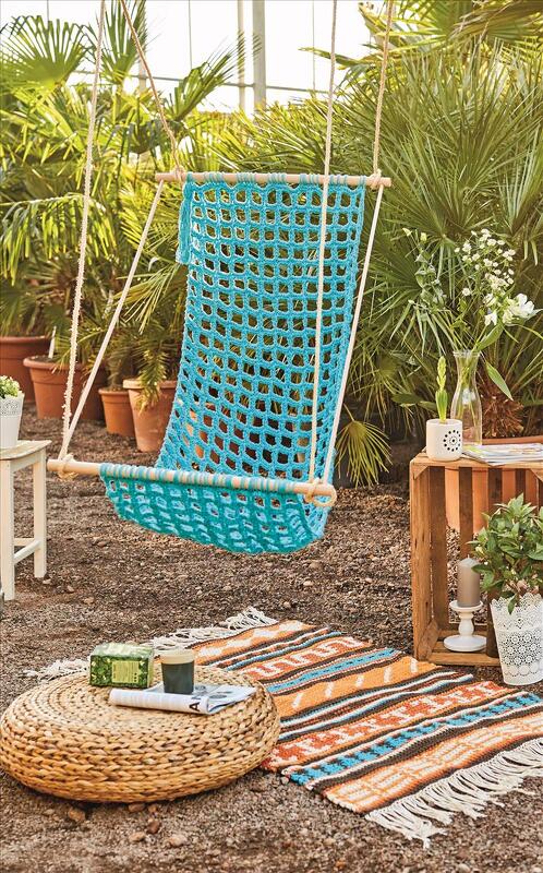 Detailreicher gehäkelter Hängesessel in Türkis mit Seilaufhängung und Holzstangen vor üppigen Pflanzen.