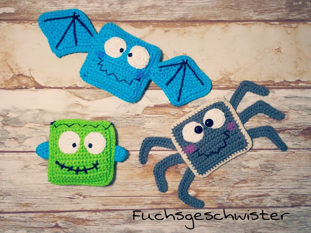 Drei gehäkelte quadratische Anhänger: blaue Fledermaus, grünes Monster und graue Spinne auf Holzuntergrund