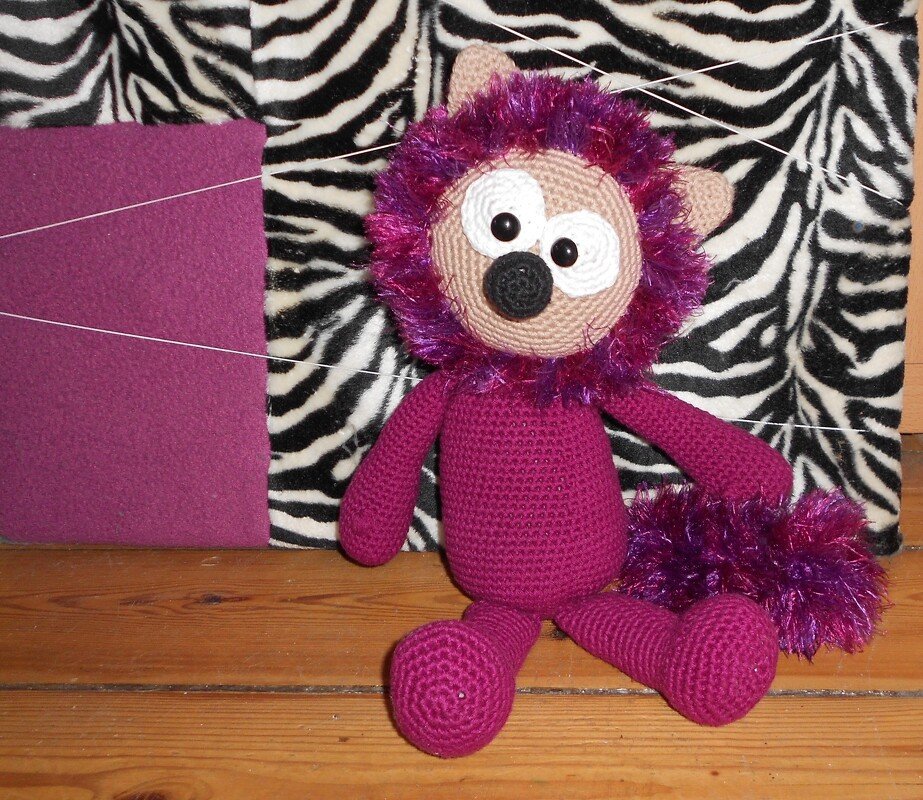 Gehäkelter lila Amigurumi-Löwe mit flauschiger violetter Mähne und buschiger Schwanzspitze vor zebraähnlichem Hintergrund