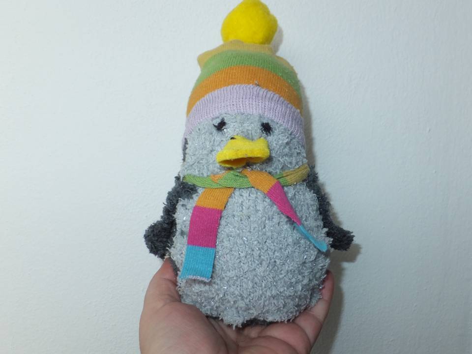 Niedlicher, grau-plüschiger Pinguin mit bunter Mütze und Schal, in einer Hand gehalten.