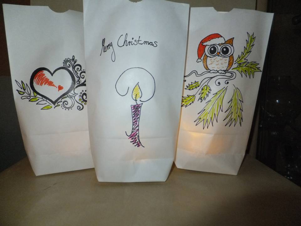 Drei beleuchtete weiße Papierbeutel mit handgemalten Weihnachtsmotiven: Herz, Kerze, Eule mit Weihnachtsmütze.