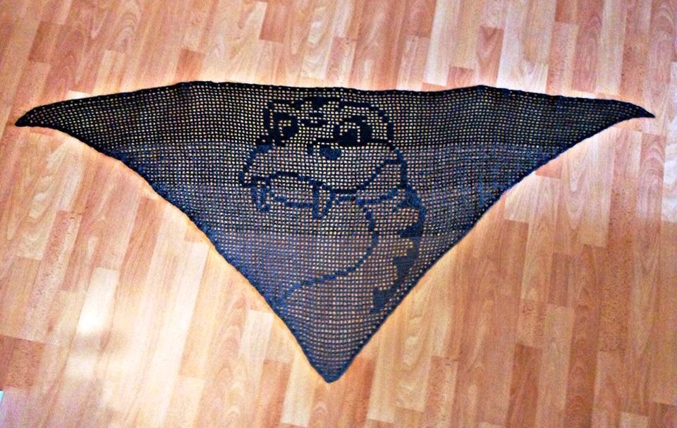 Dunkles, dreieckiges Häkel­tuch mit eingearbeitetem Monster‑Motiv auf hellem Holzboden.