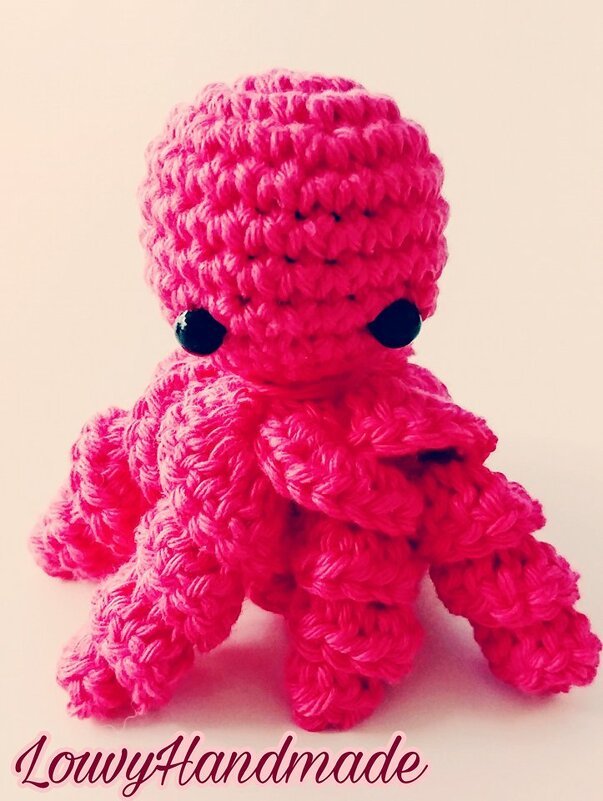 Vorderansicht eines kleinen pinken gehäkelten Amigurumi‑Oktopus mit schwarzen Augen auf hellem Hintergrund.