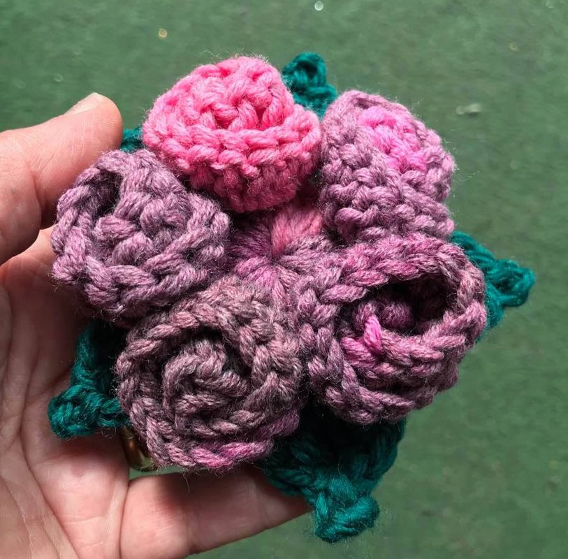 Nahaufnahme mehrerer zarter Häkelrosen in Rosatönen mit grünen Blättern in einer Hand