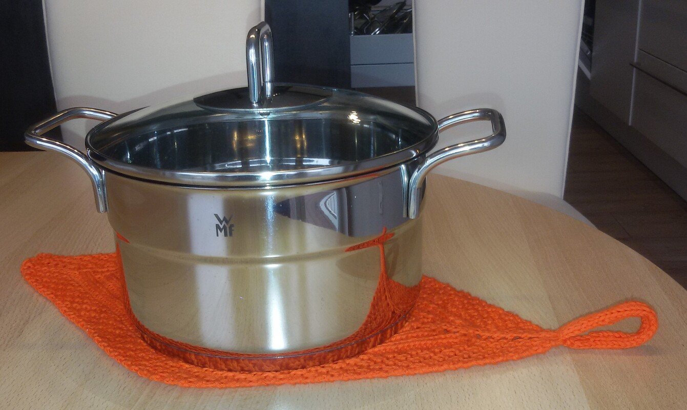 Orangefarbener gestrickter Topflappen mit Aufhängeschlaufe unter einem Kochtopf auf Holztisch.